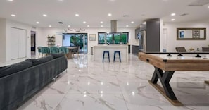 A modern living room with a pool table, a sofa, and a kitchen area in the background.