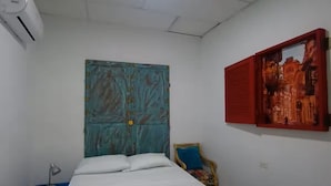 Ein Schlafzimmer mit einem Bett, einem blauen Kopfteil, einer Wand-Klimaanlage, einem Sessel mit Kissen und einem gerahmten Bild an der Wand.