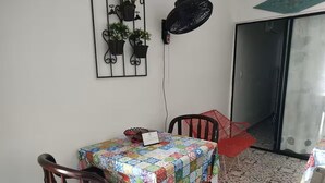 Ein Essbereich mit Tisch, der mit einer farbenfrohen Decke bedeckt ist, zwei Stühle, ein roter Sessel und ein Wandventilator.