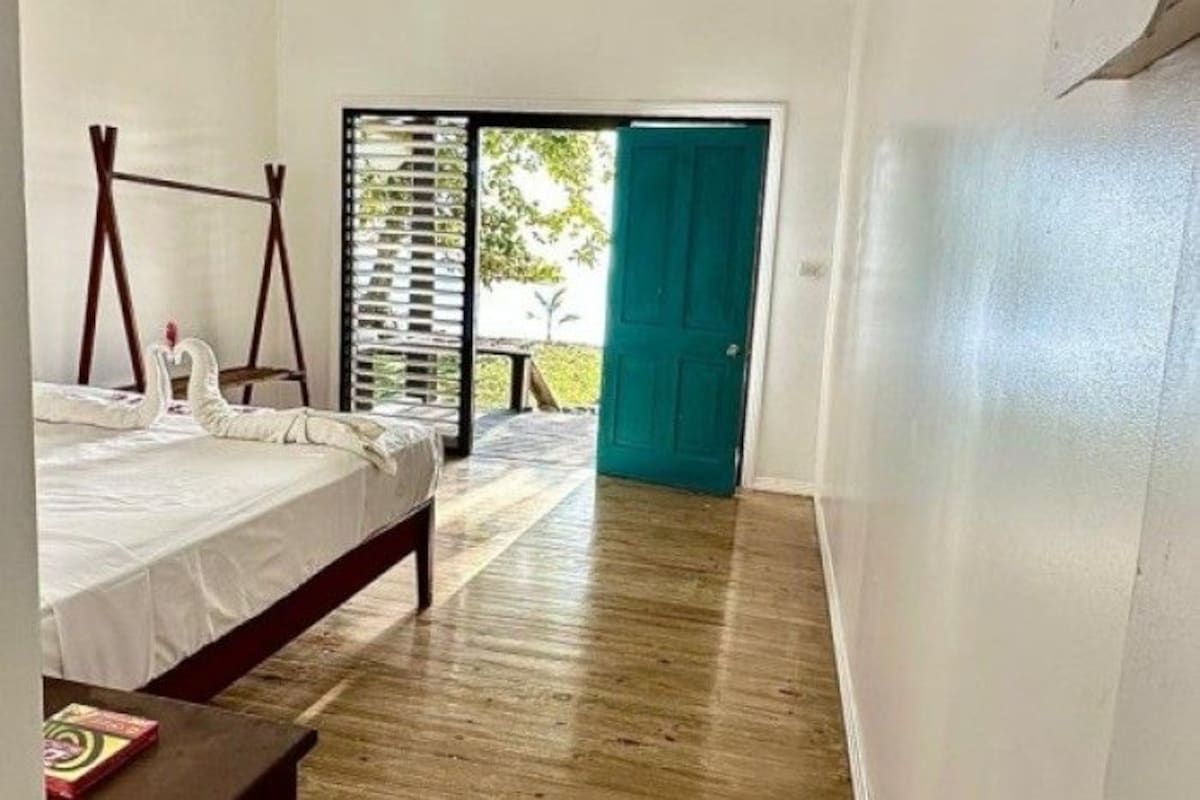 A bedroom with a bed, a wooden floor, a green door, and a view through a window.