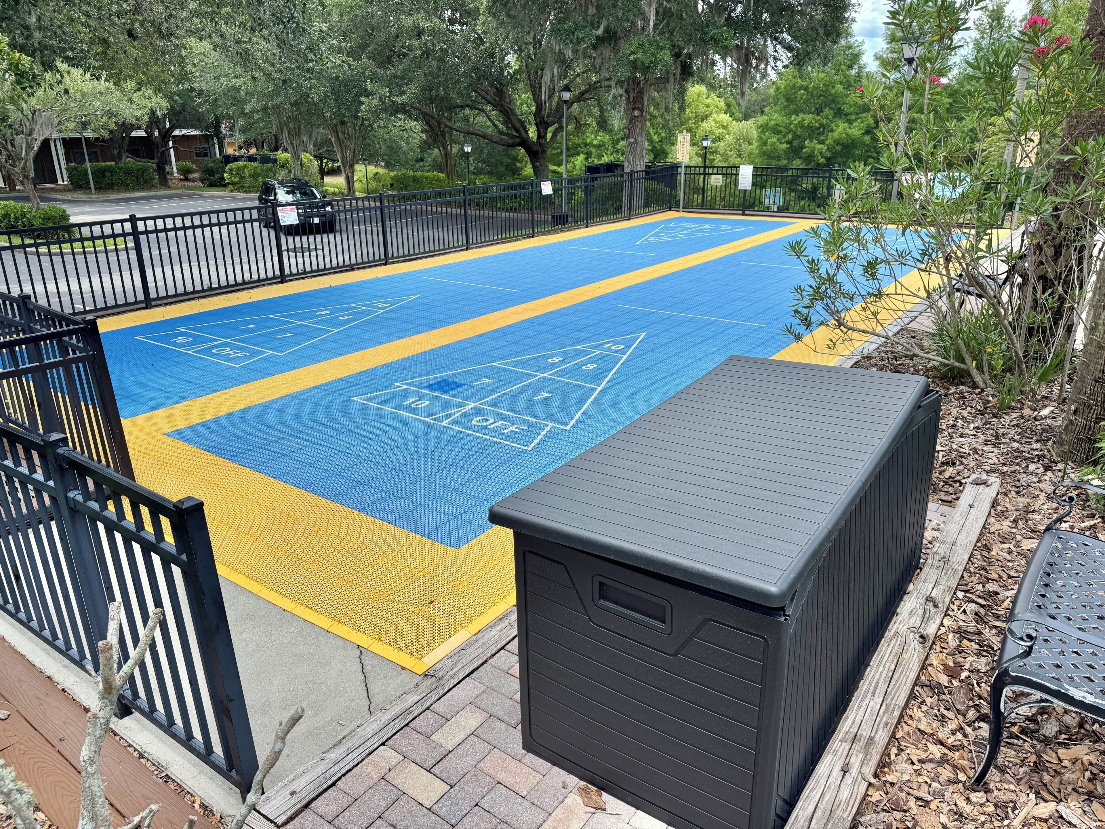 Ein blaues und gelbes Shuffleboardfeld mit einem schwarzen Mülleimer und einer Bank in der Nähe.