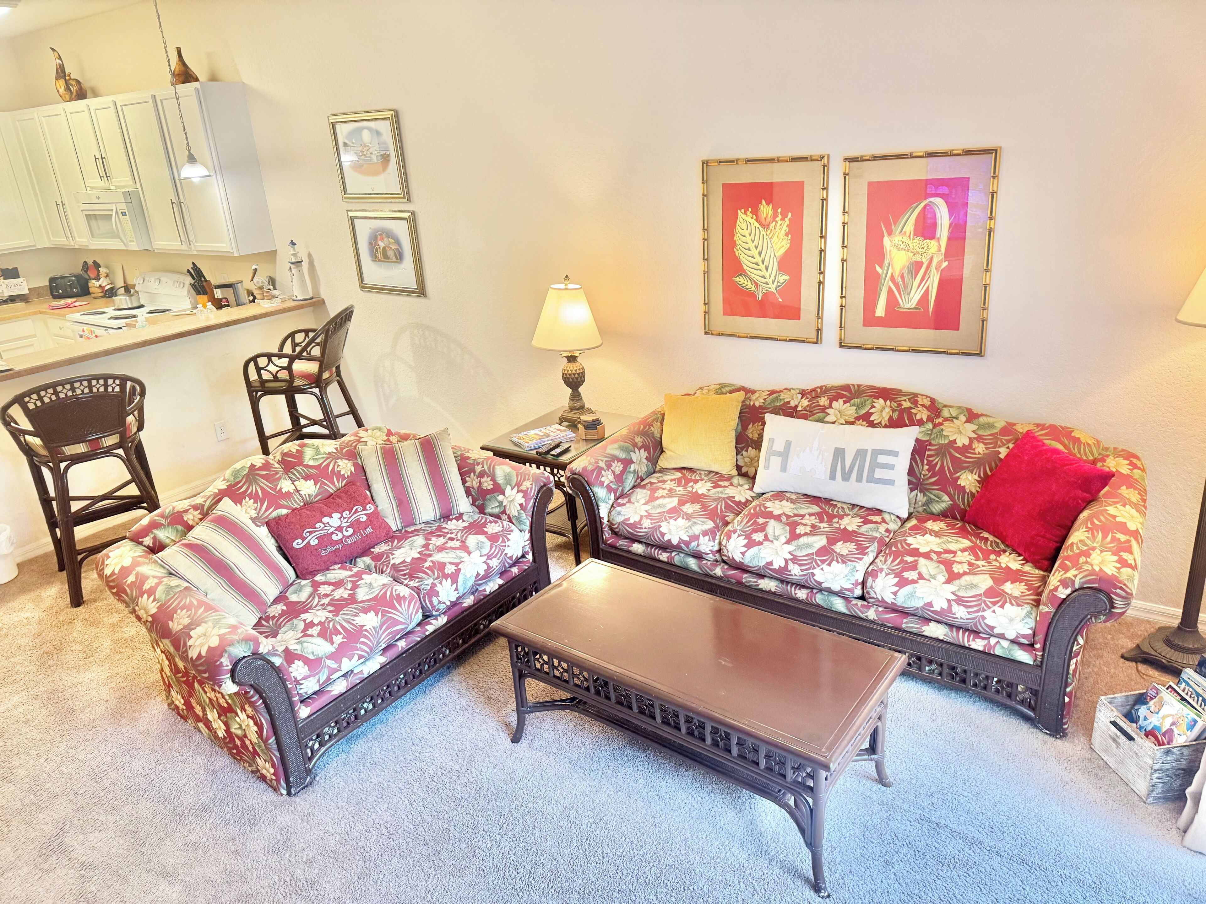 Un salon avec un canapé à motifs floraux, un fauteuil assorti et une table basse. Il y a également un coin cuisine avec des meubles blancs et un four à micro-ondes.