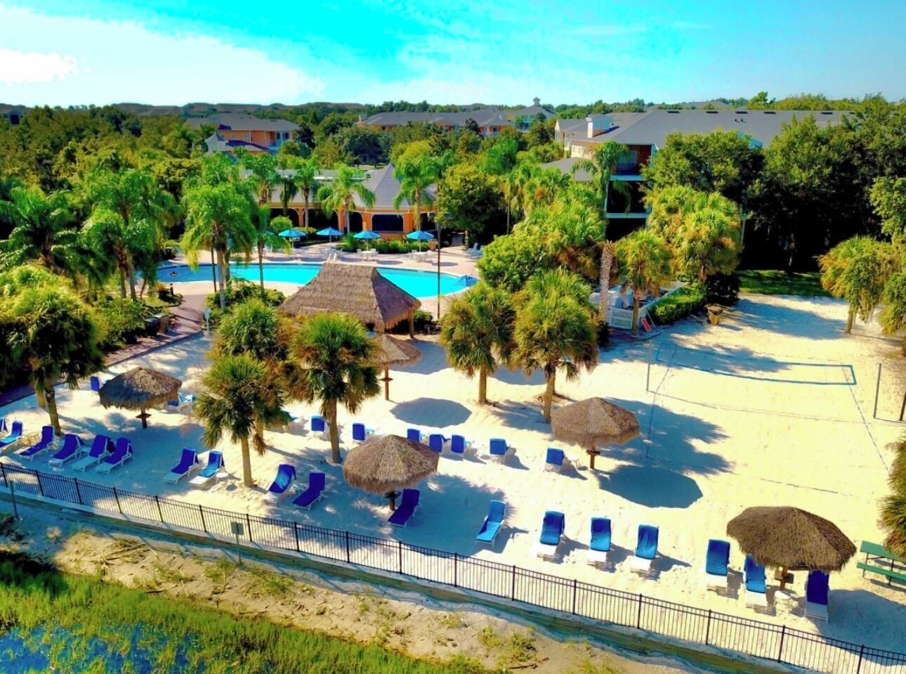 Een resort met een zwembad, ligbedden, palmbomen en een omheind strand.