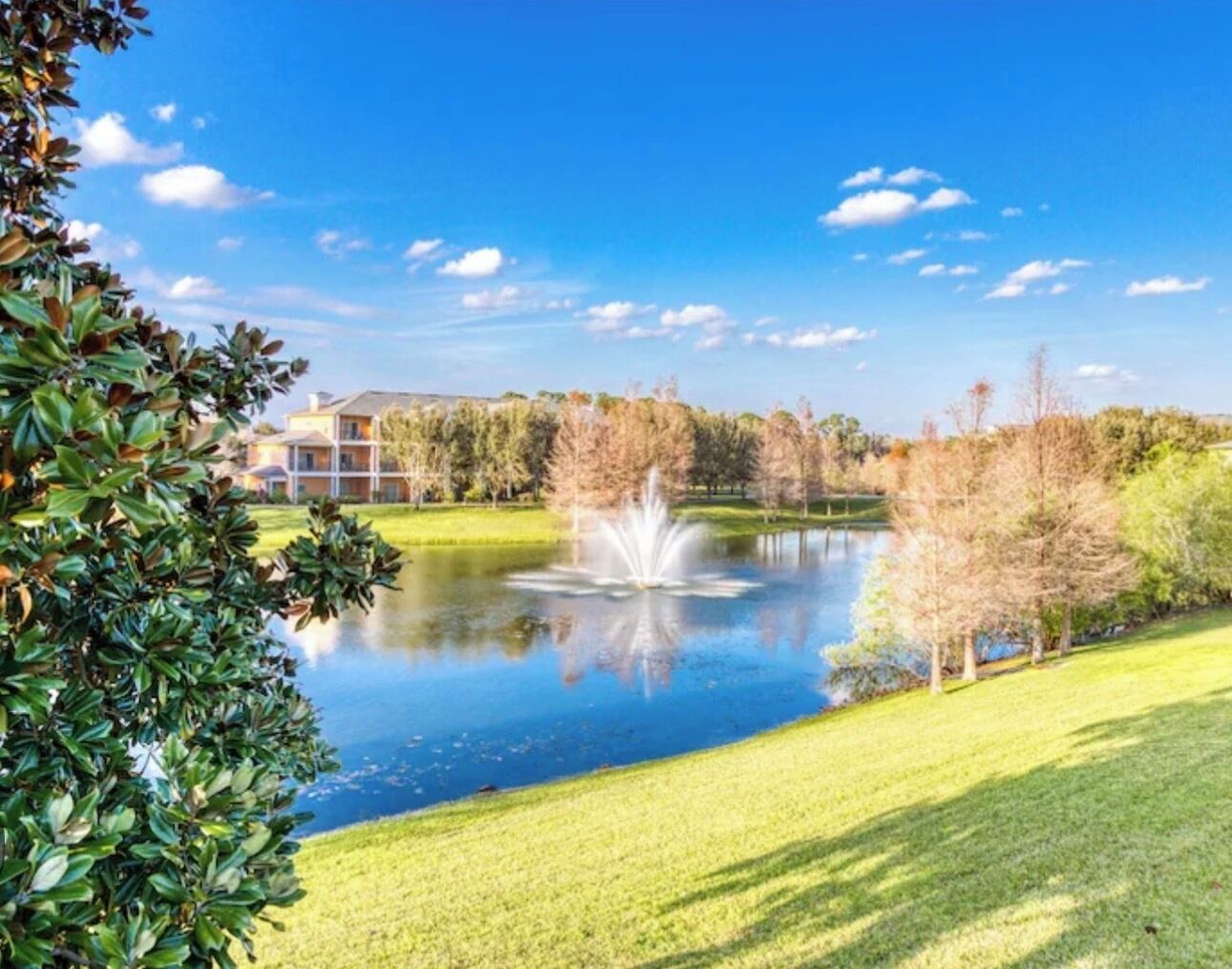 A serene lake with a fountain, surrounded by well-maintained grass and trees, with a building in the background.