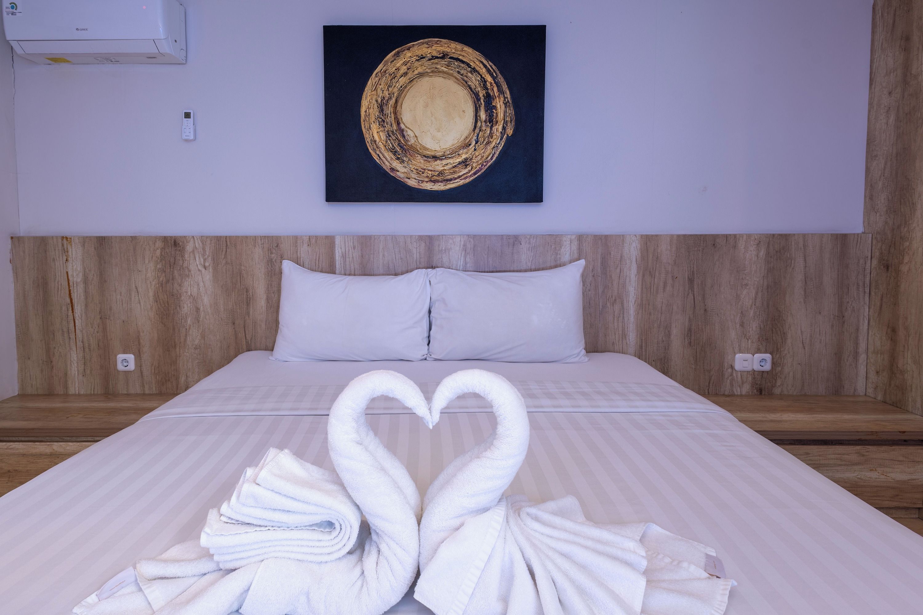 Une chambre d’hôtel avec un lit, un climatiseur fixé au mur, un tableau et une serviette pliée en forme de cœur.