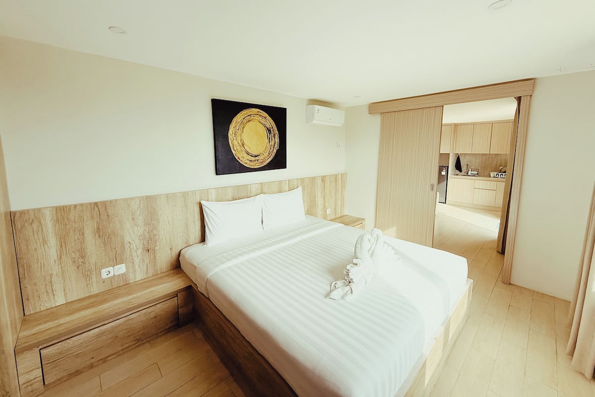 A hotel room with a bed, a wooden headboard, a wall-mounted air conditioner, and a view into the kitchen area.
