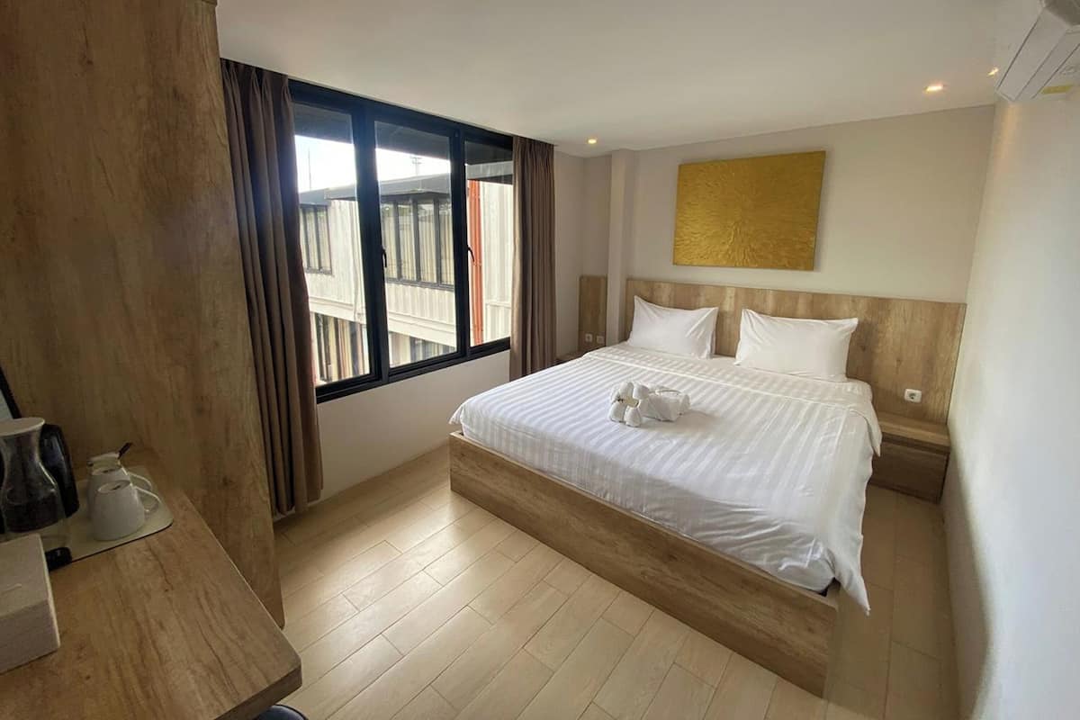 A hotel room with a large bed, a wooden headboard, a window with curtains, a wall-mounted artwork, and a small table with a kettle and cups.