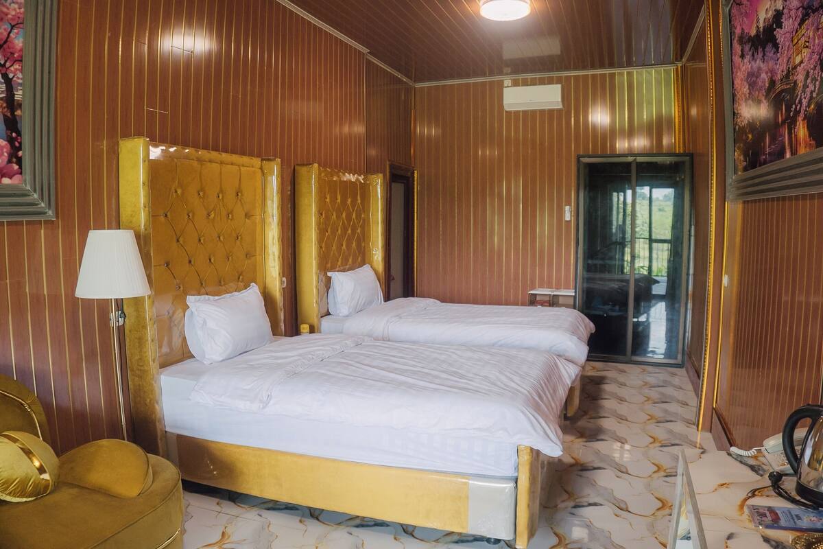 Quarto de hotel com duas camas, cabeceira dourada, poltrona dourada, luminária e vista para o exterior pela janela.