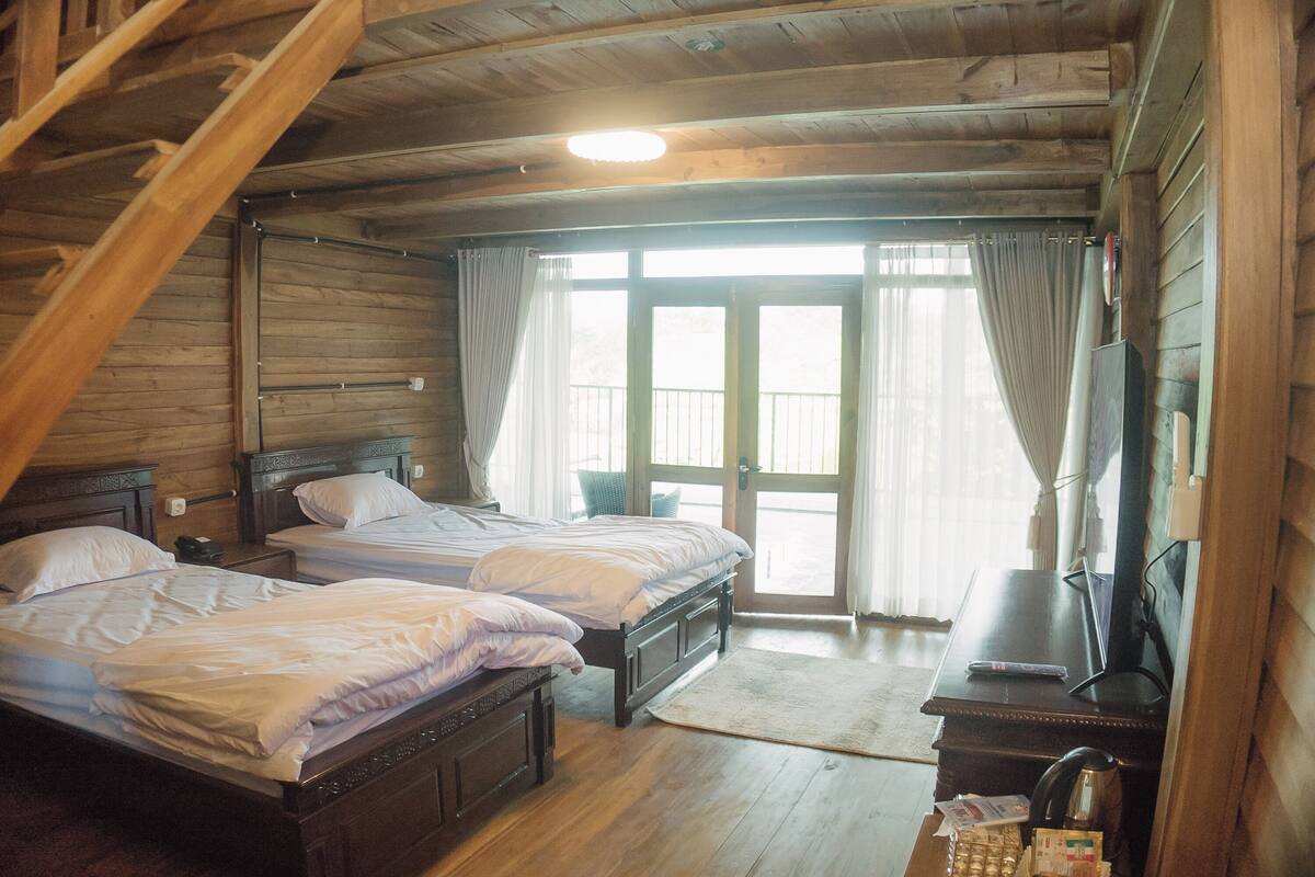 Um quarto com duas camas de madeira, uma escrivaninha com cadeira, televisão e uma janela com cortinas.