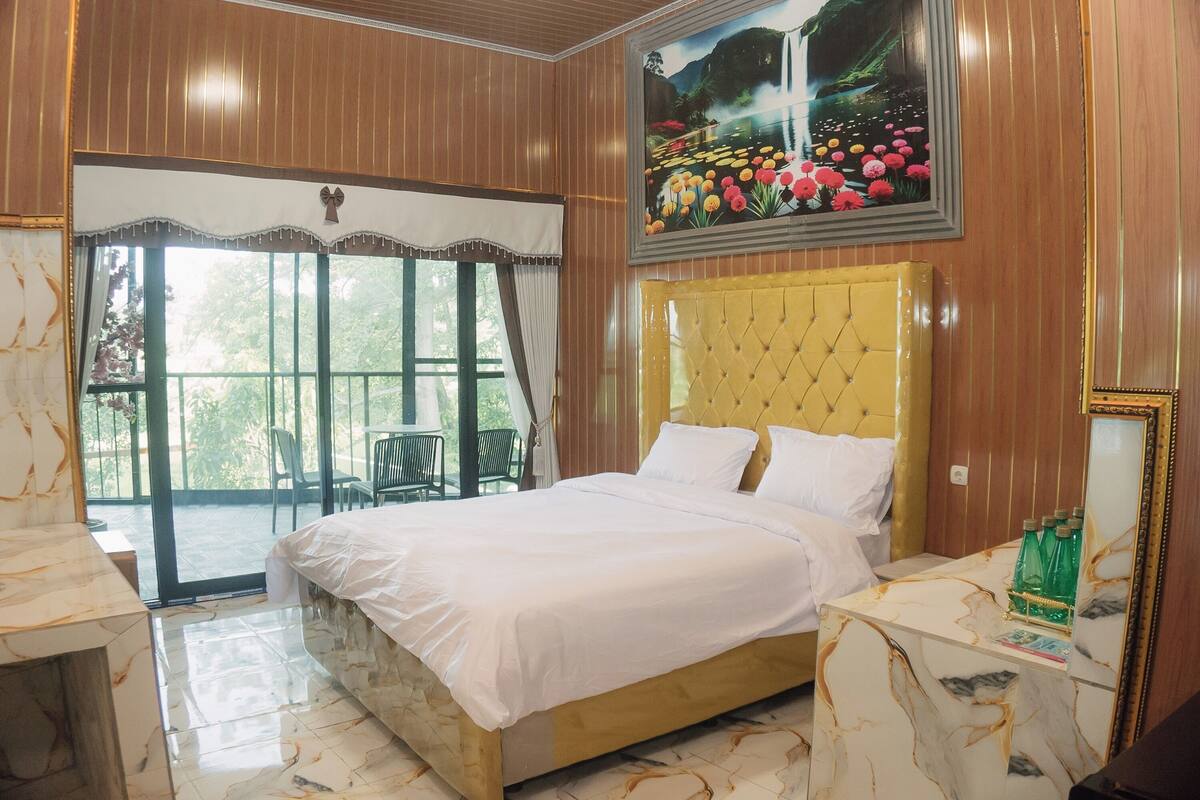 Quarto com cama, mesa de cabeceira de mármore, janela grande com cortinas e pintura de flores.