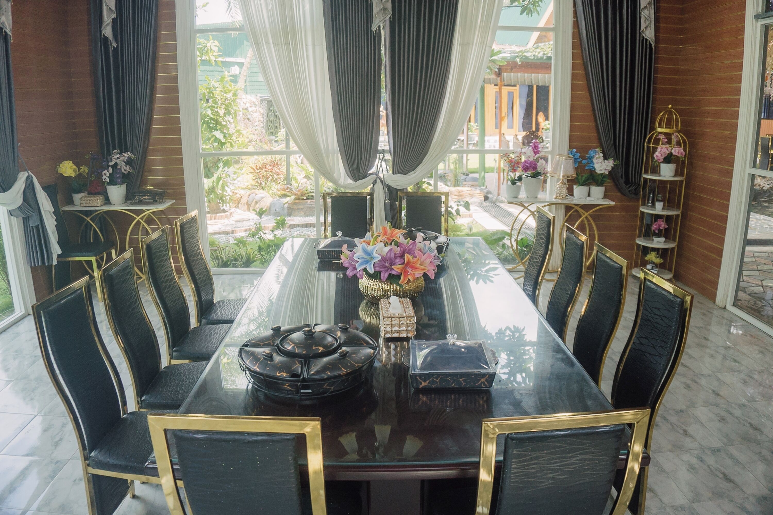 A dining area with a glass table, black chairs with gold frames, and a view of the garden through large windows.