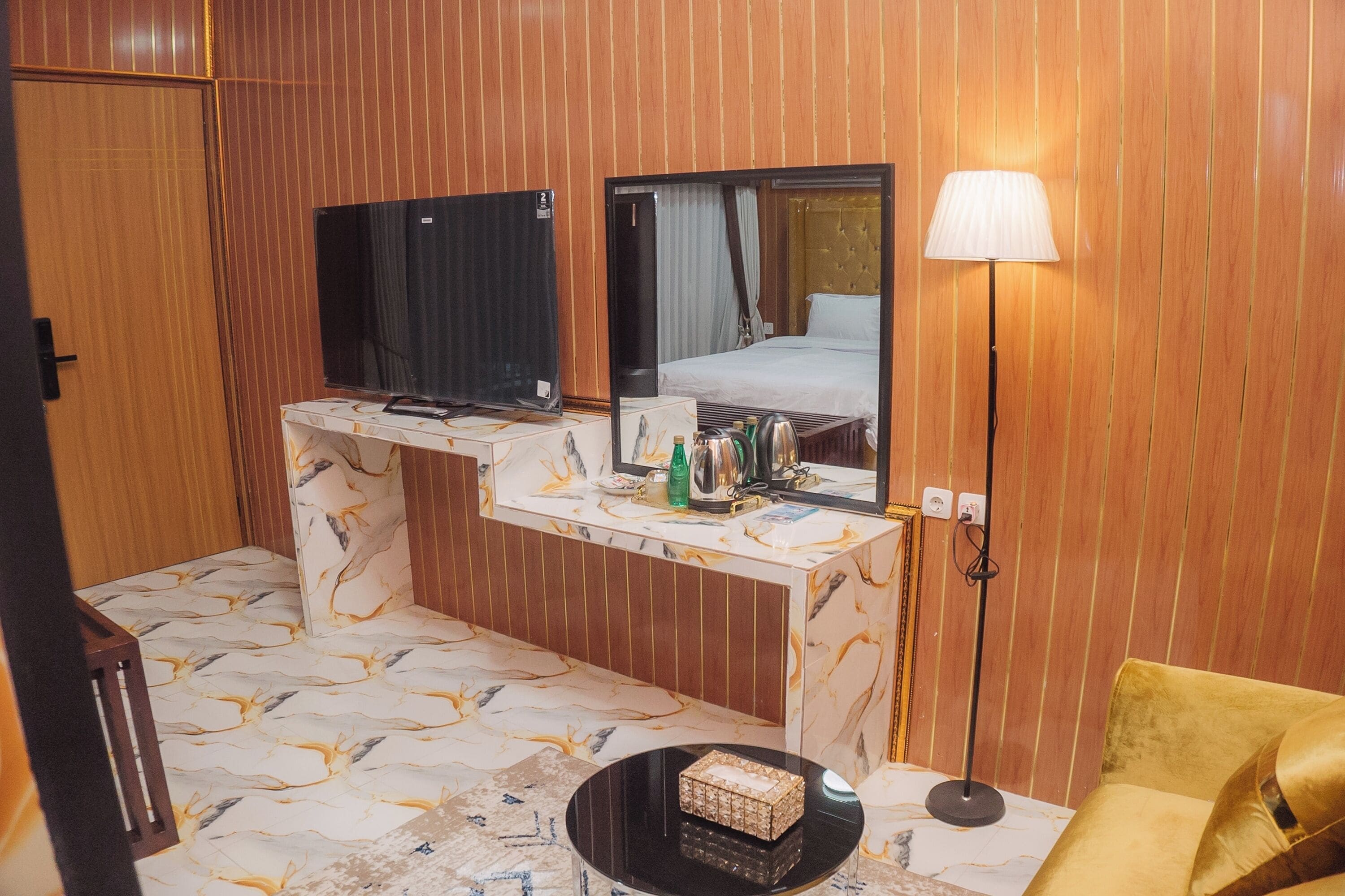 A hotel room with a bed, a TV, a desk with a lamp, and a small table with a box.
