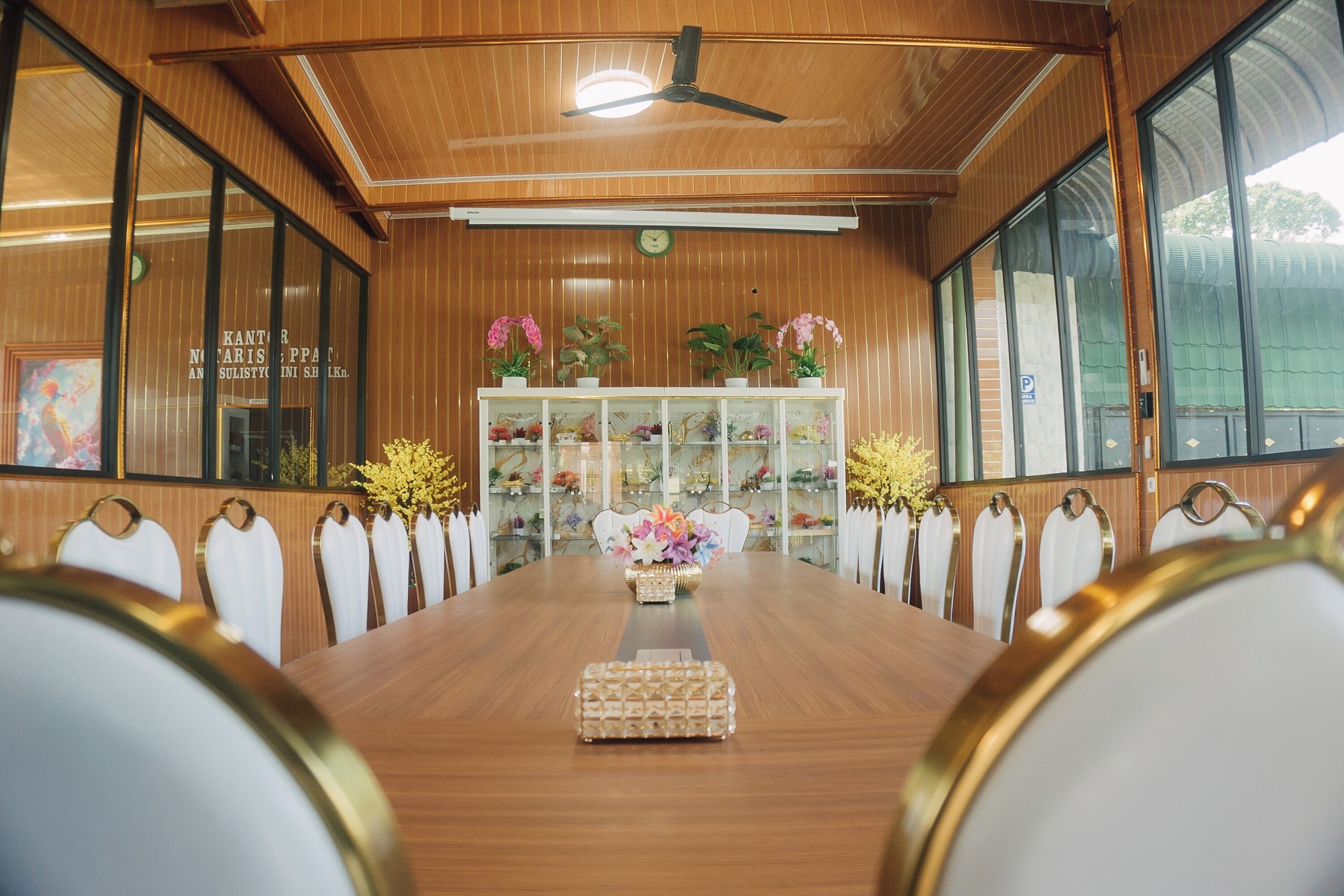 A conference room with a long wooden table, white chairs, a ceiling fan, and a display cabinet with plants.
