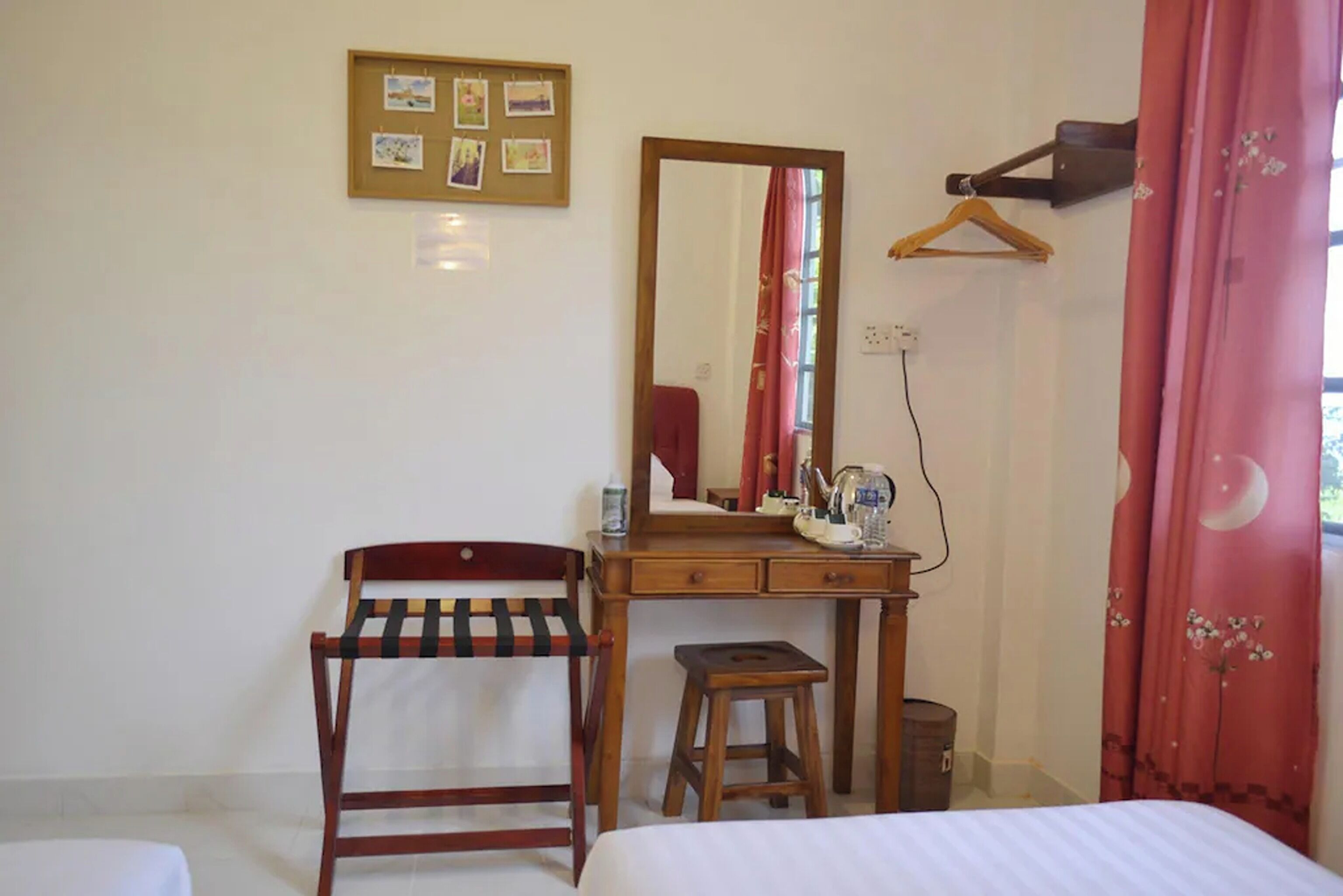 Una habitación con una cama, un escritorio de madera con un espejo, una silla, un taburete pequeño y un perchero en la pared.