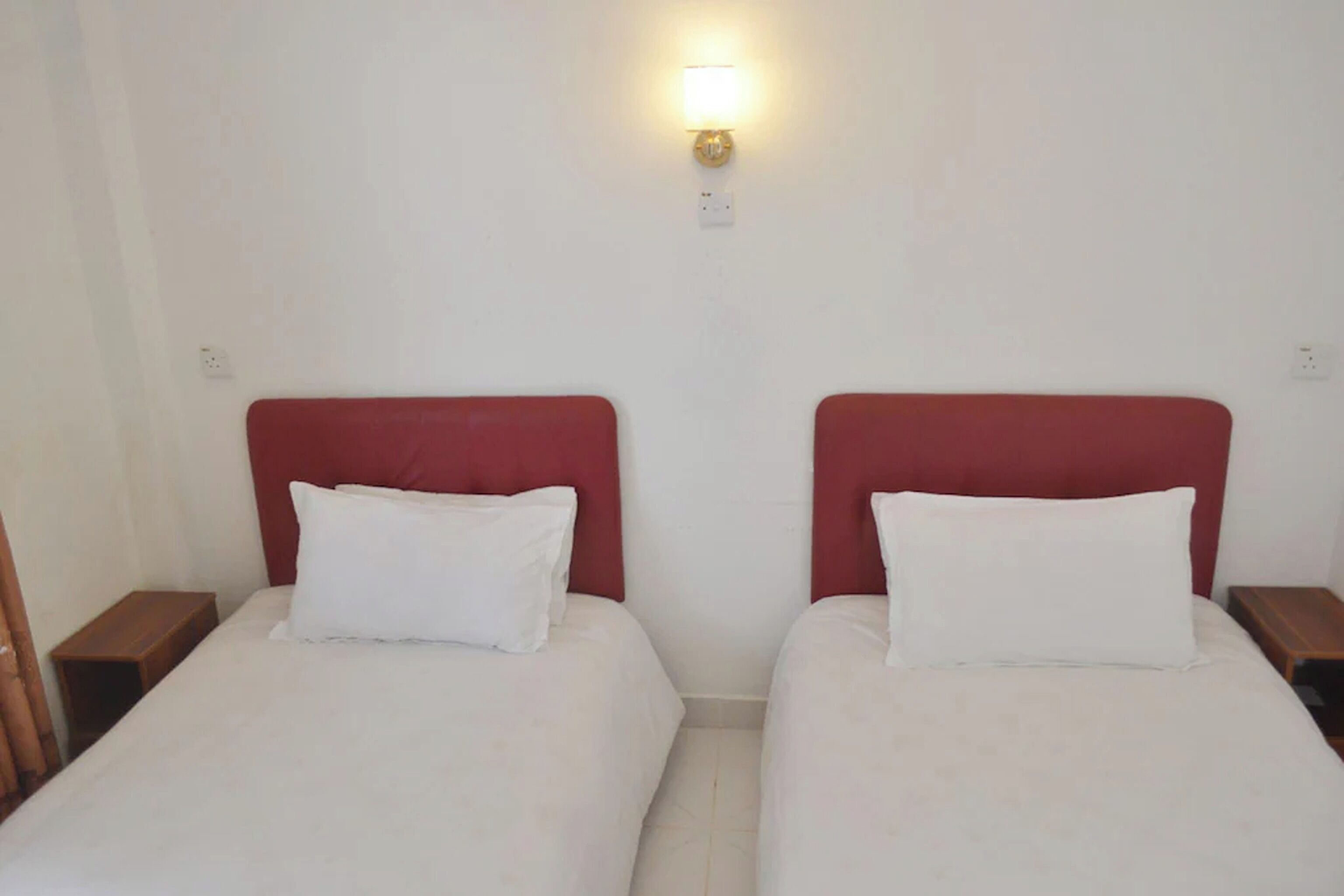 A hotel room with two beds, each with a red headboard and white bedding. There is a wall-mounted light fixture above the beds.