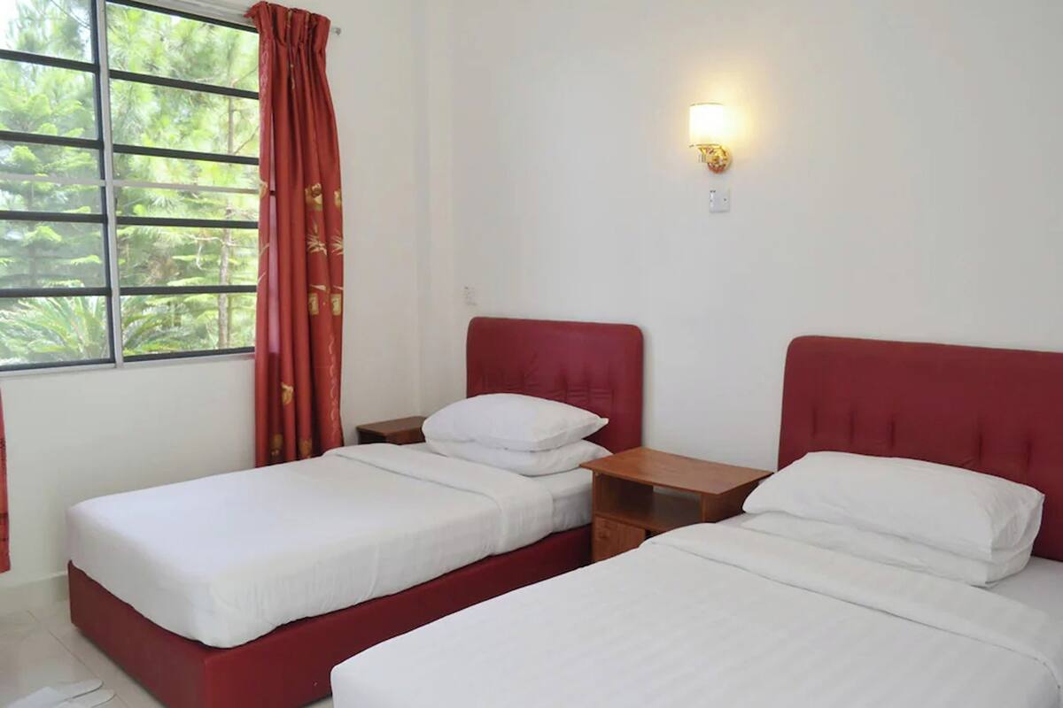 A room with two single beds, a window with red curtains, a nightstand, and a wall-mounted light.