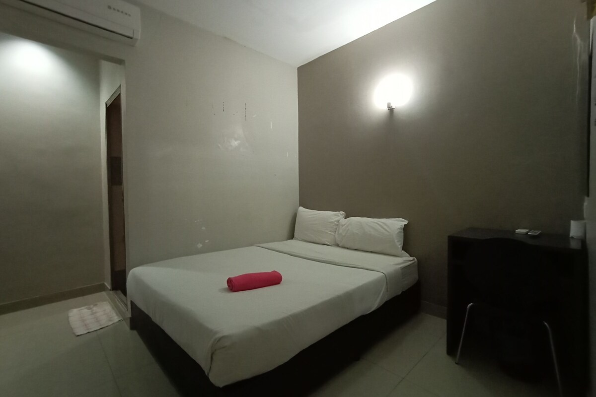 A small, simple room with a bed, a desk, and an air conditioning unit.