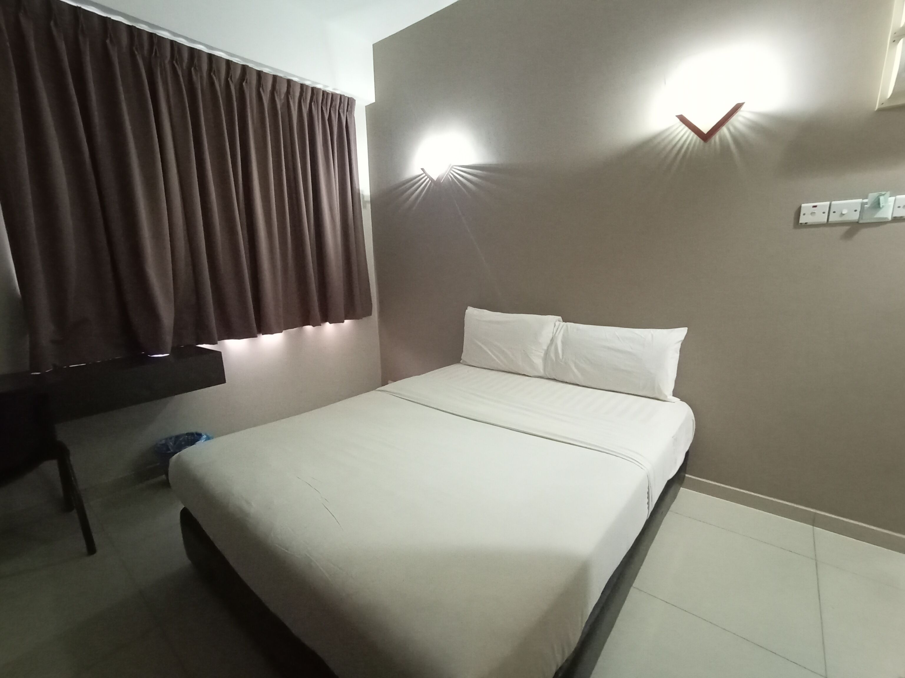 Une chambre d’hôtel avec un lit, des rideaux, un bureau et des lampes fixées au mur.
