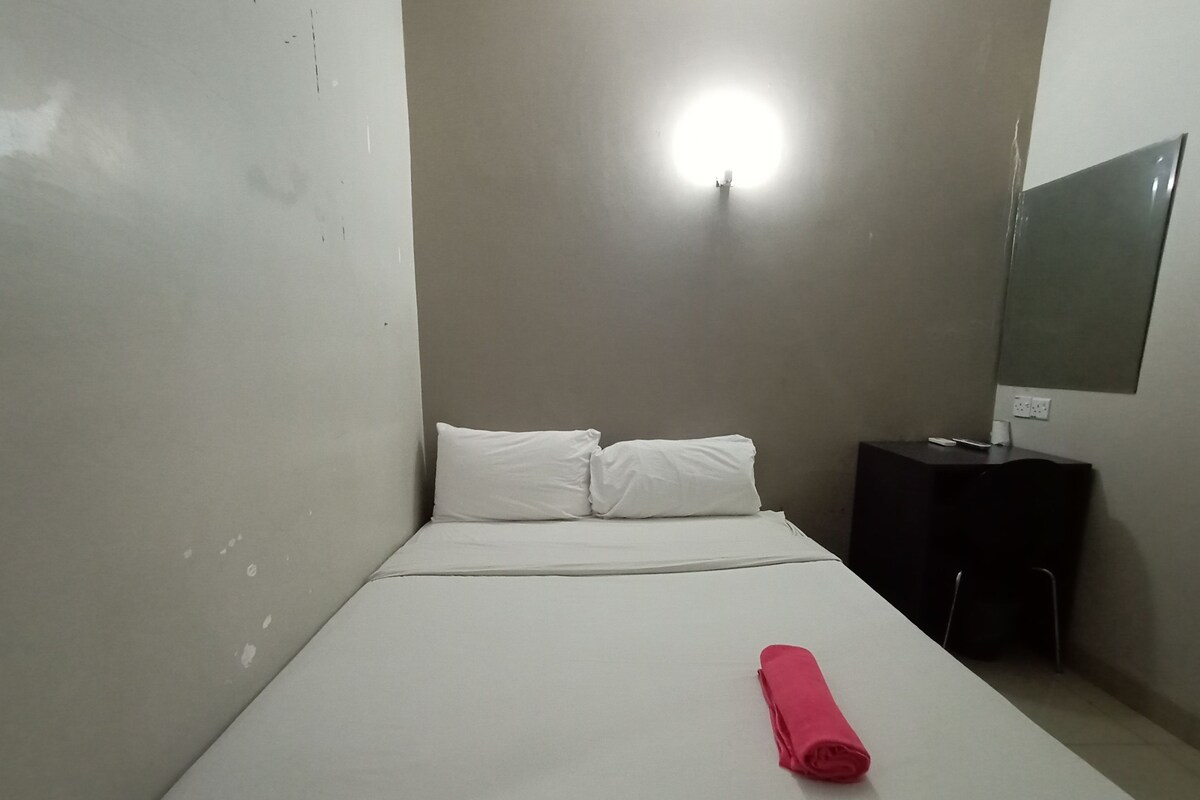 A small, single-bed room with a white bed, a small bedside table, a mirror, and a wall-mounted light.