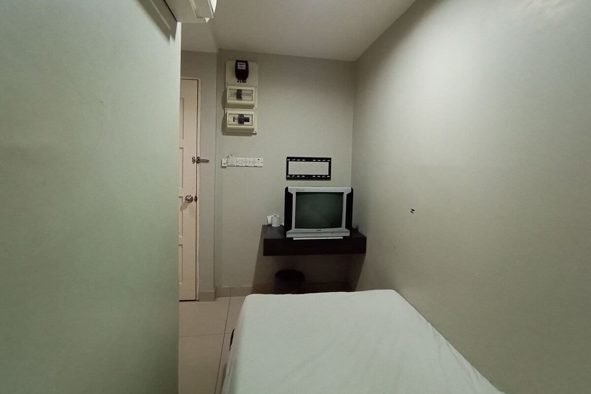 A small, dimly lit room with a bed, a television, and a door leading to another room.