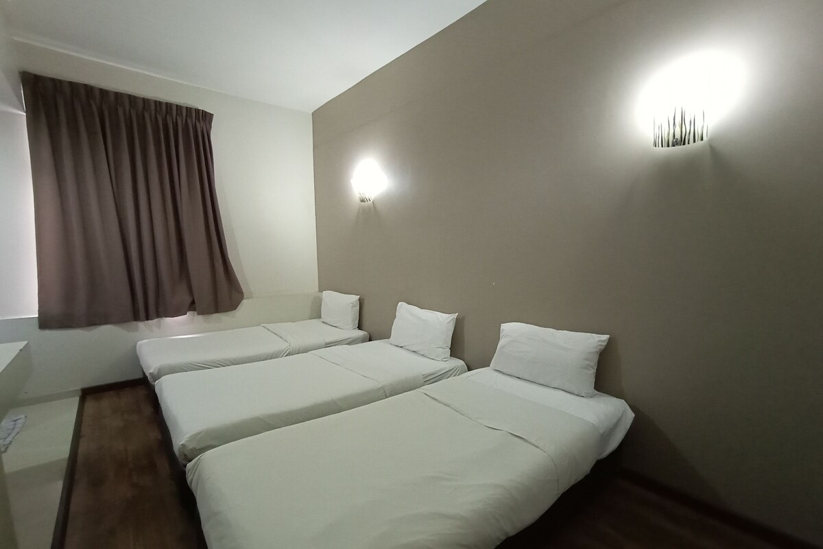 A hotel room with two single beds, a window with curtains, a bedside table, and a wall-mounted light fixture.