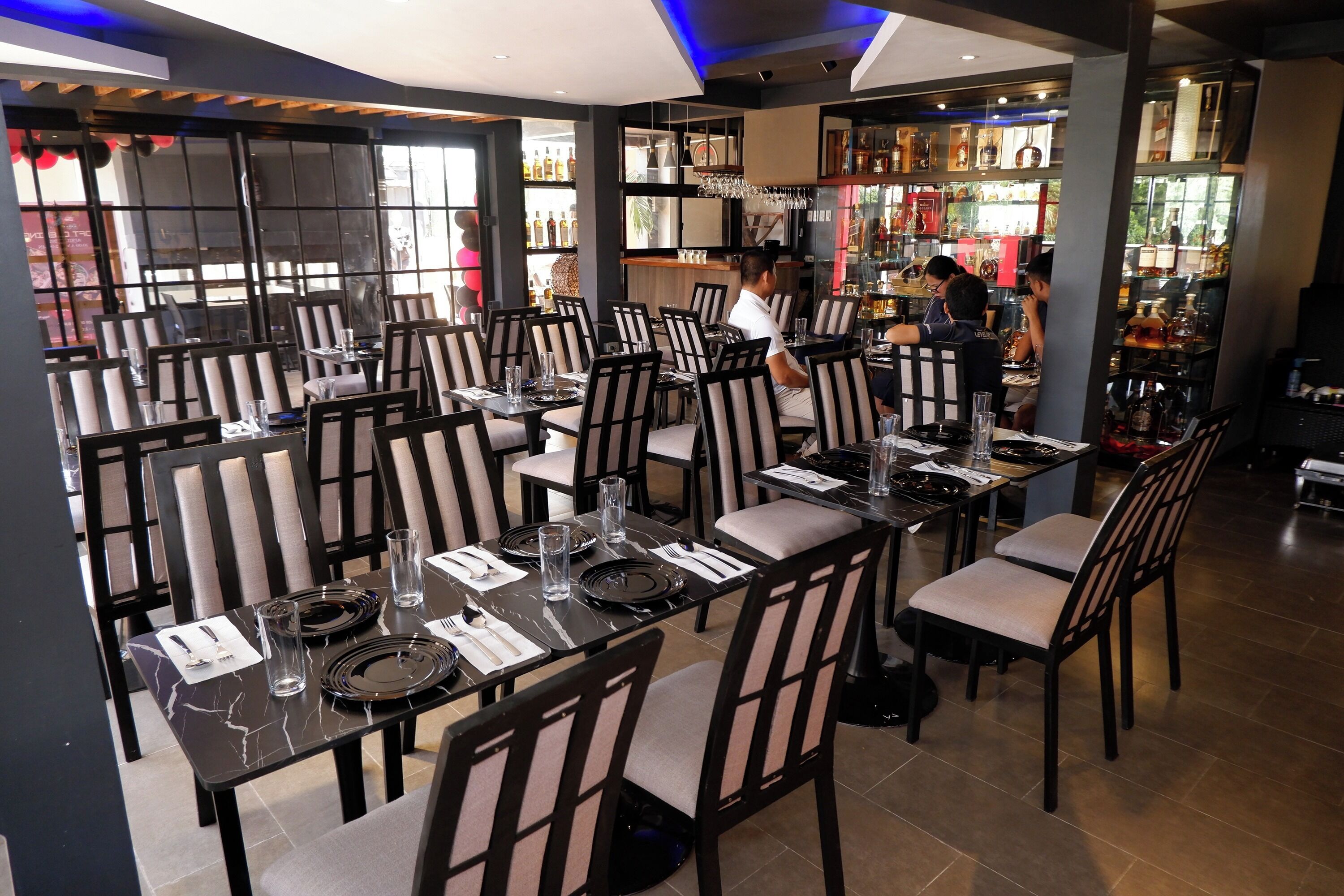 A restaurant with multiple tables set for dining, a bar area with shelves of bottles, and a view through a large window.