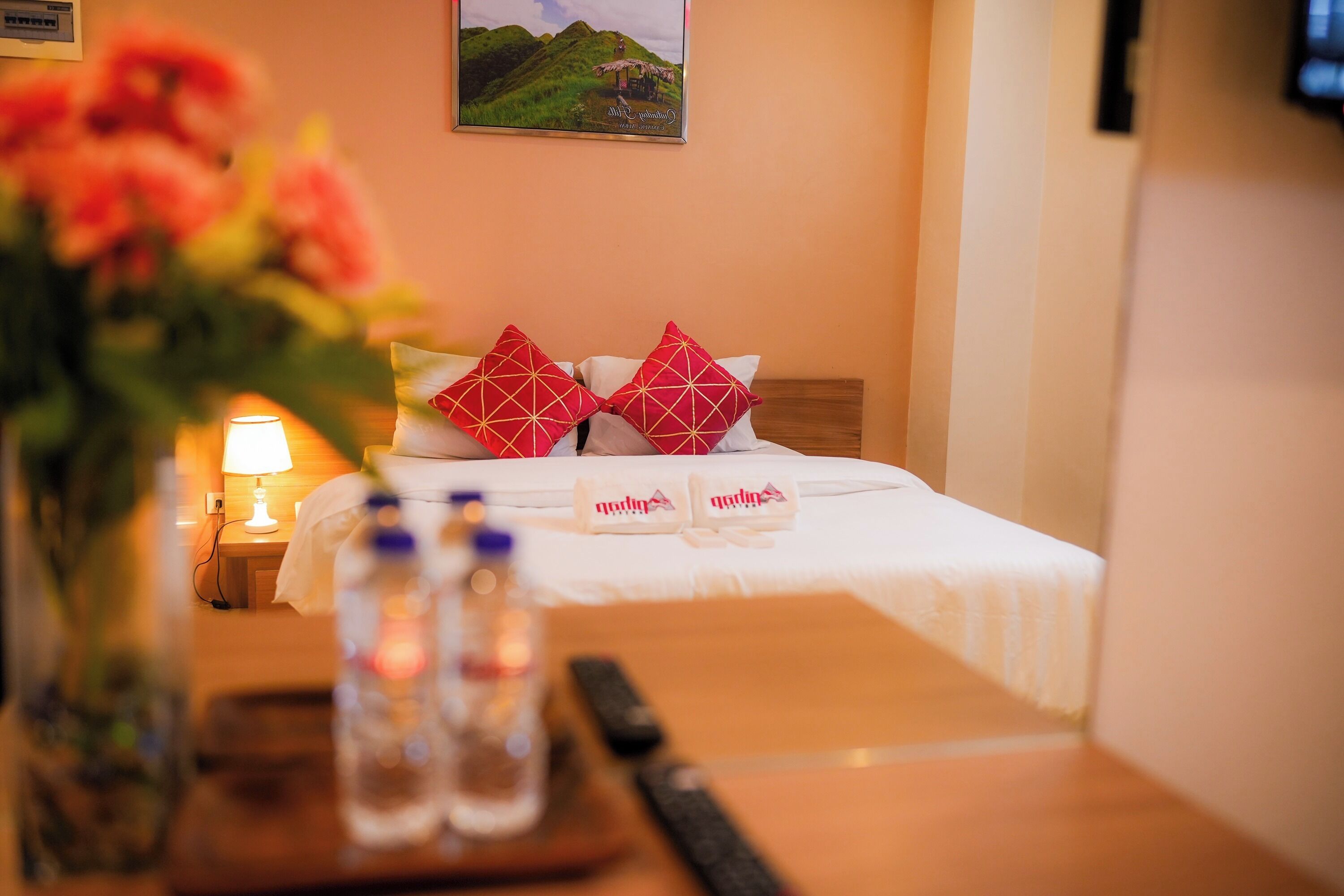Habitación de hotel con una cama, almohadas, una mesita de noche con lámpara, un florero con flores y un cuadro en la pared.