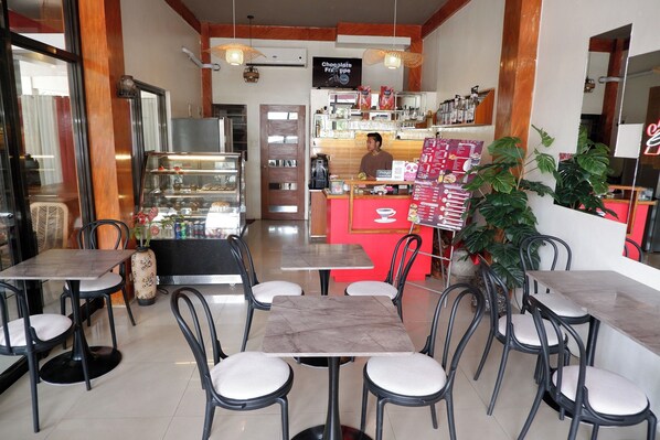 Un café avec des tables et des chaises surmontées de plateaux en marbre, un comptoir où est affiché le menu, et une vitrine présentant divers produits au chocolat.