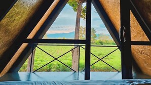 Una habitación con techo inclinado, una cama y vistas a un paisaje verde a través de una ventana con barandilla de metal.