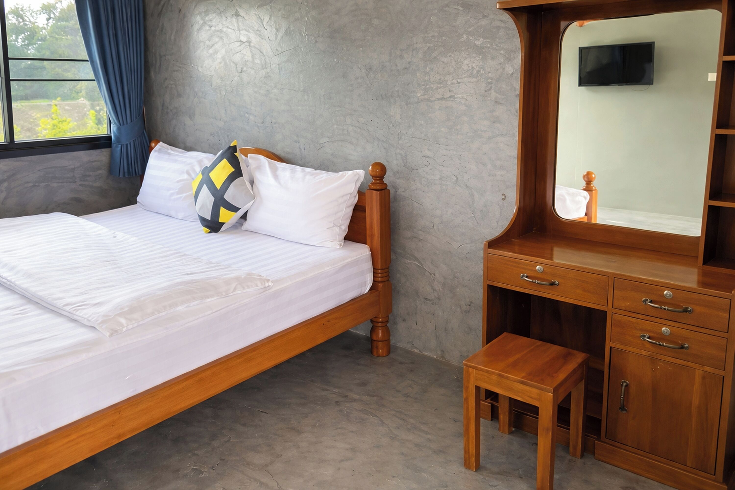 Une chambre à coucher avec un lit en bois, un petit tabouret en bois, une commode en bois avec des tiroirs et un téléviseur fixé au mur.