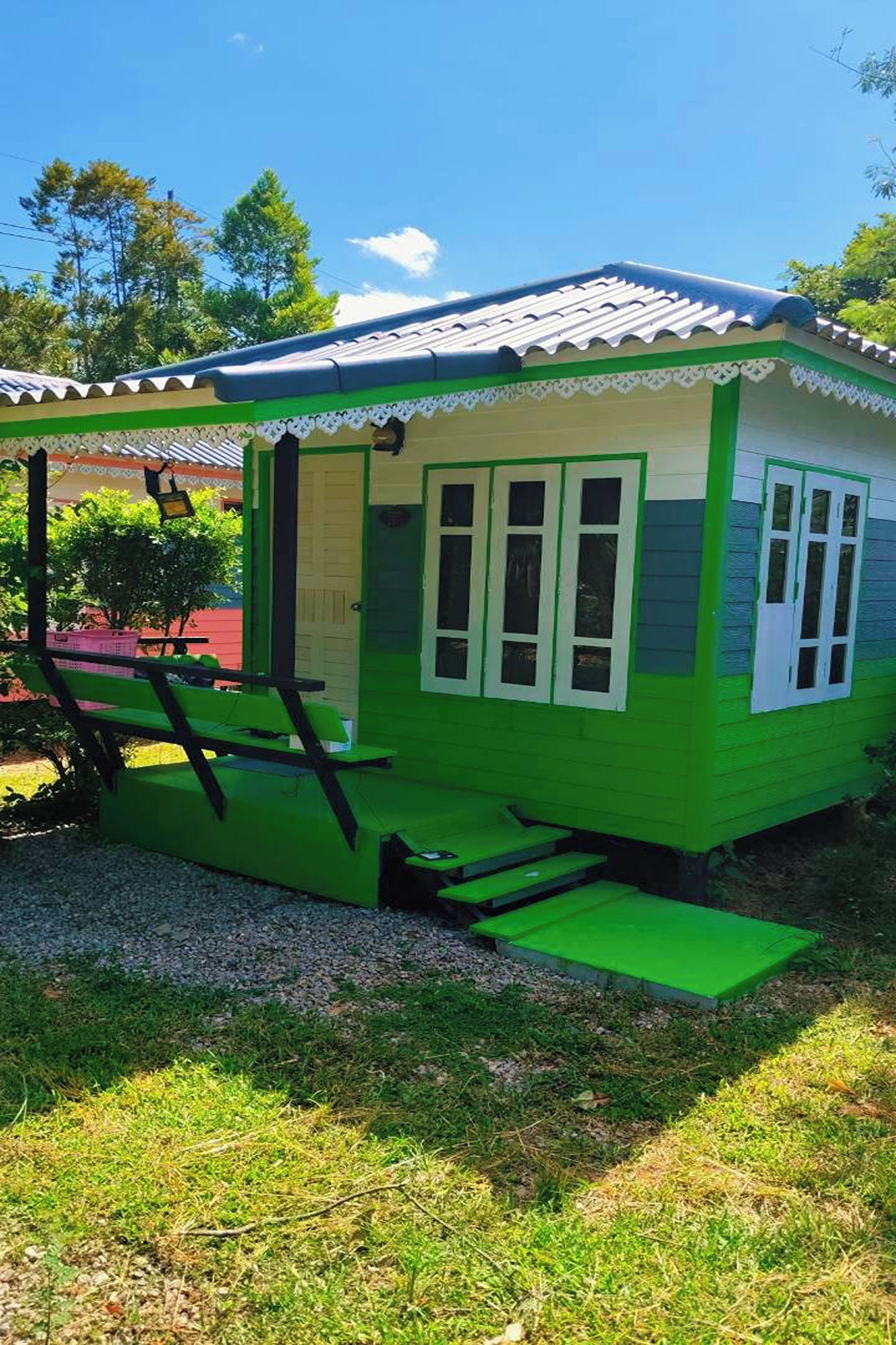 Une petite maison à un seul étage, à l’extérieur vert et aux bordures blanches, dotée d’un porche avec un banc et une petite table.