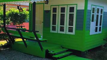 Une petite maison à un seul étage, à l’extérieur vert et aux bordures blanches, dotée d’un porche avec un banc et une petite table.