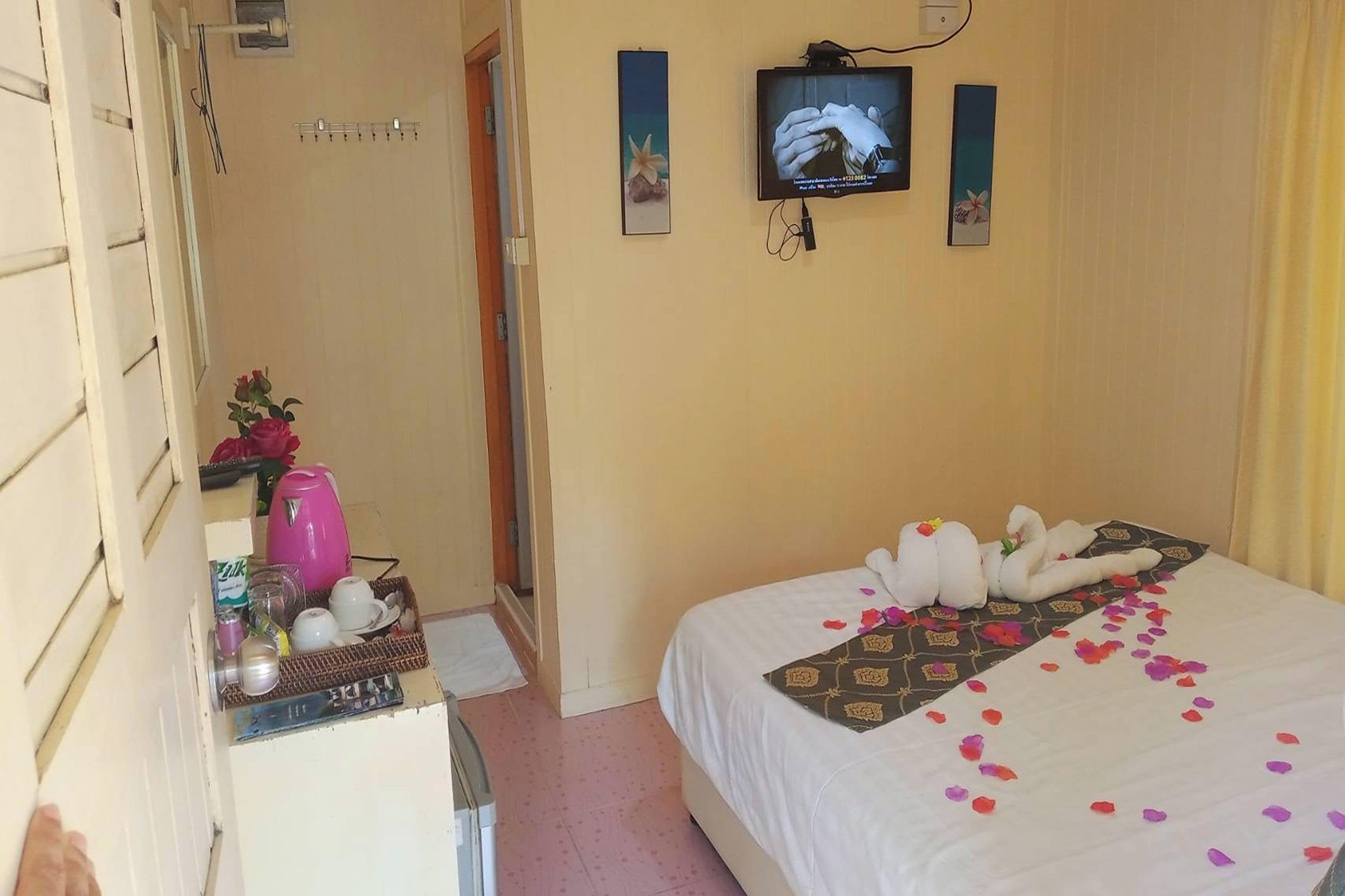 A hotel room with a bed, a television, a small table with a pink kettle, and a door leading to another room.