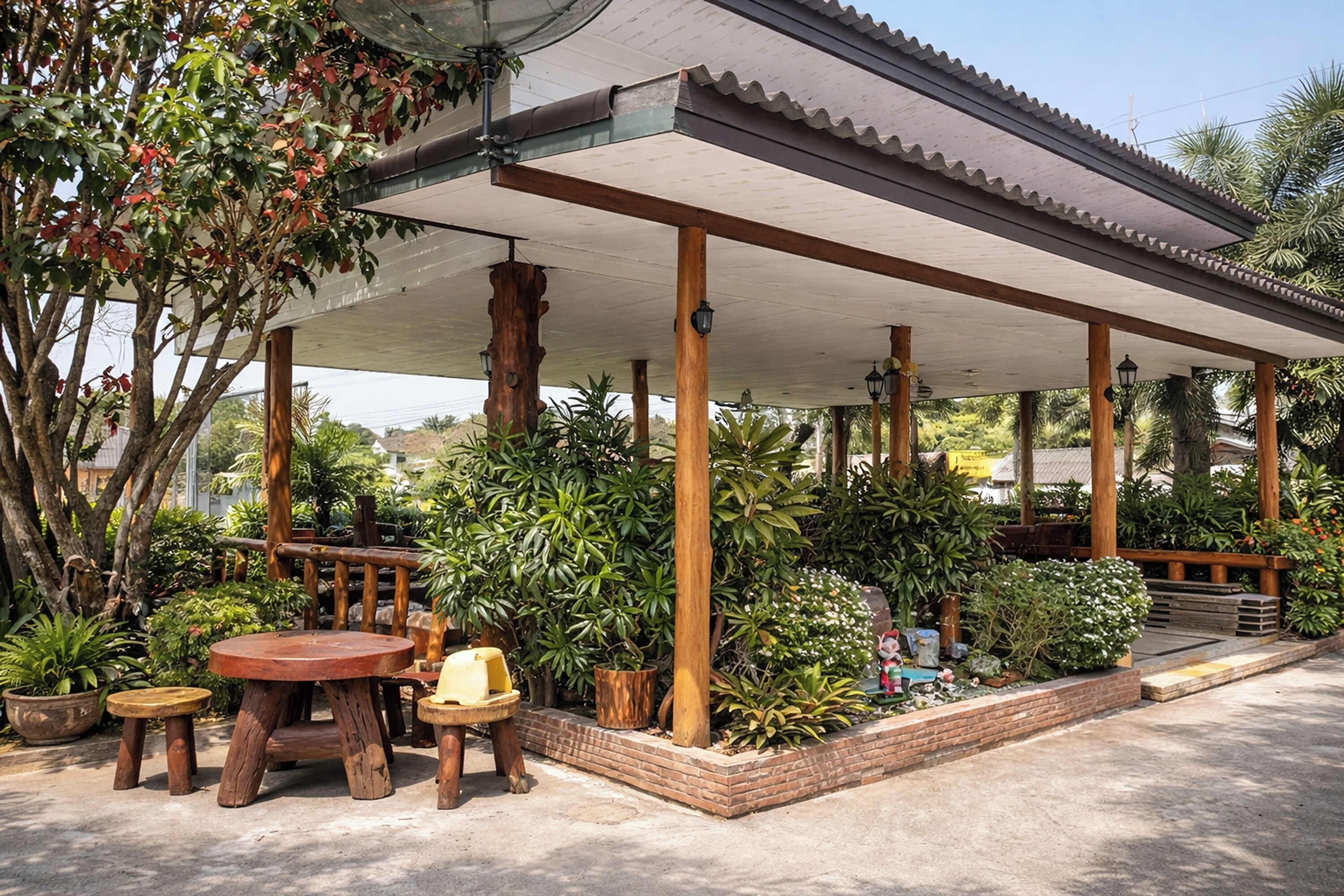 A covered outdoor seating area with wooden tables and chairs, surrounded by potted plants and trees.
