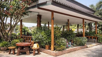 A covered outdoor seating area with wooden tables and chairs, surrounded by potted plants and trees.