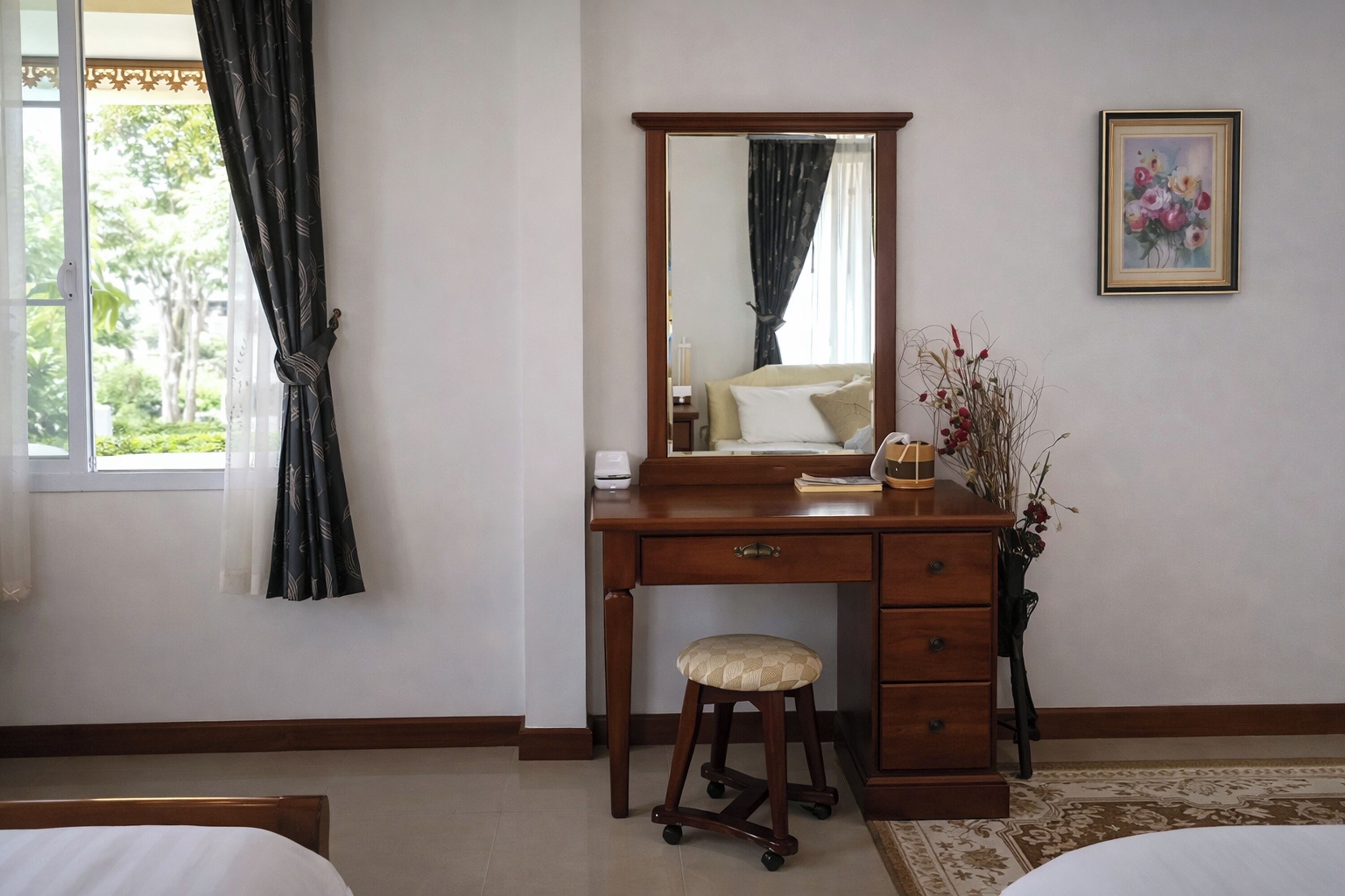 A bedroom with a wooden desk, a mirror, a bedside table, a bed, and a window with curtains.