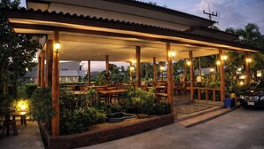 Ristorante all'aperto con tavoli e sedie in legno, immerso nel verde e dotato di un'area coperta illuminata.