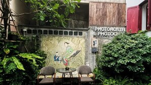 Um pequeno espaço ao ar livre com duas cadeiras e uma mesa, uma parede com um mural e uma placa com a inscrição 'PHOTOHOSTEL & PHOTO'.