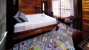 Quarto com teto de madeira, uma cama com travesseiros, uma cadeira, uma mesa e um chão colorido revestido de azulejos.