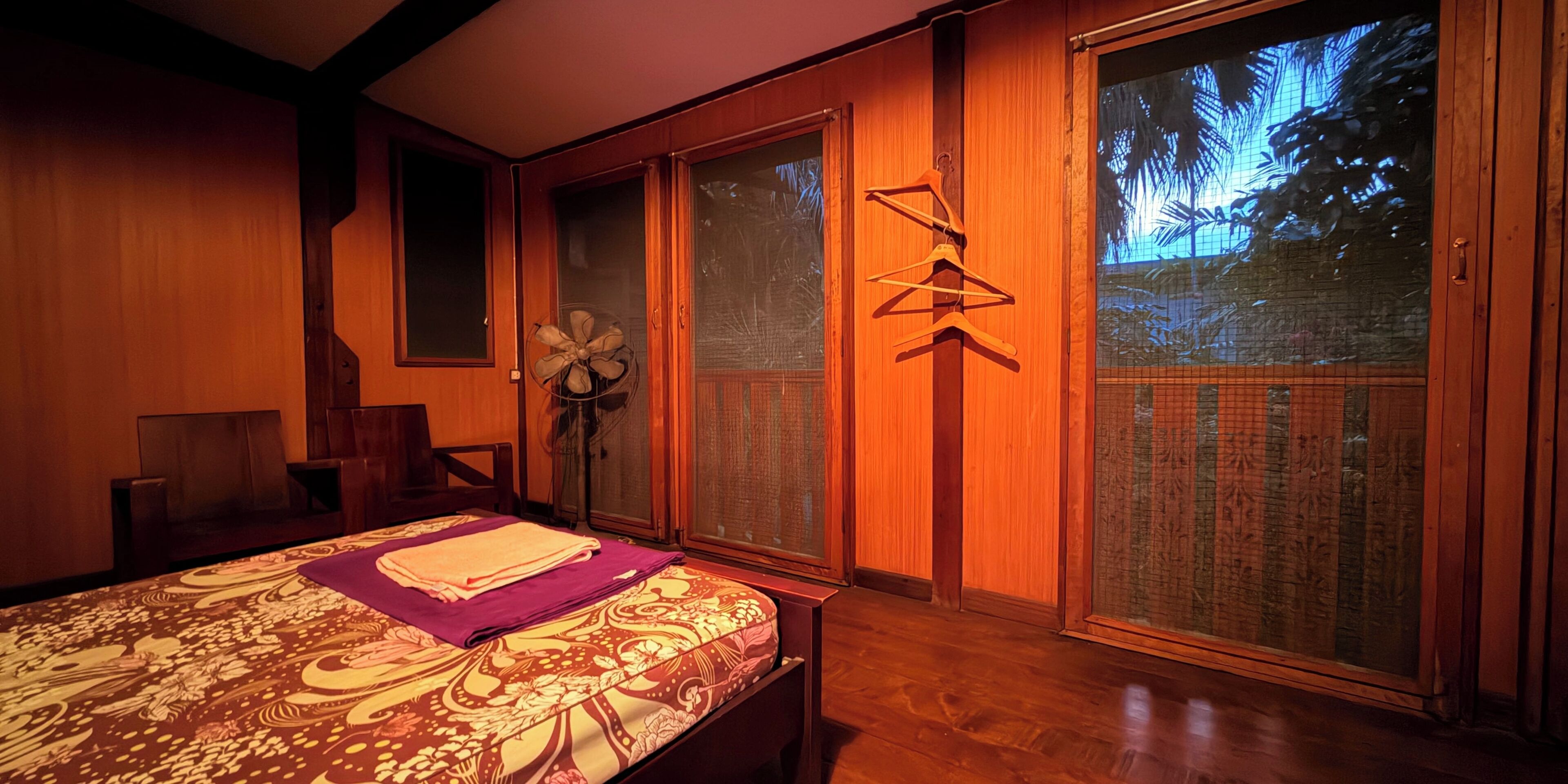 A room with a bed, a fan, a wooden cabinet, and a sliding glass door with a view of palm trees.