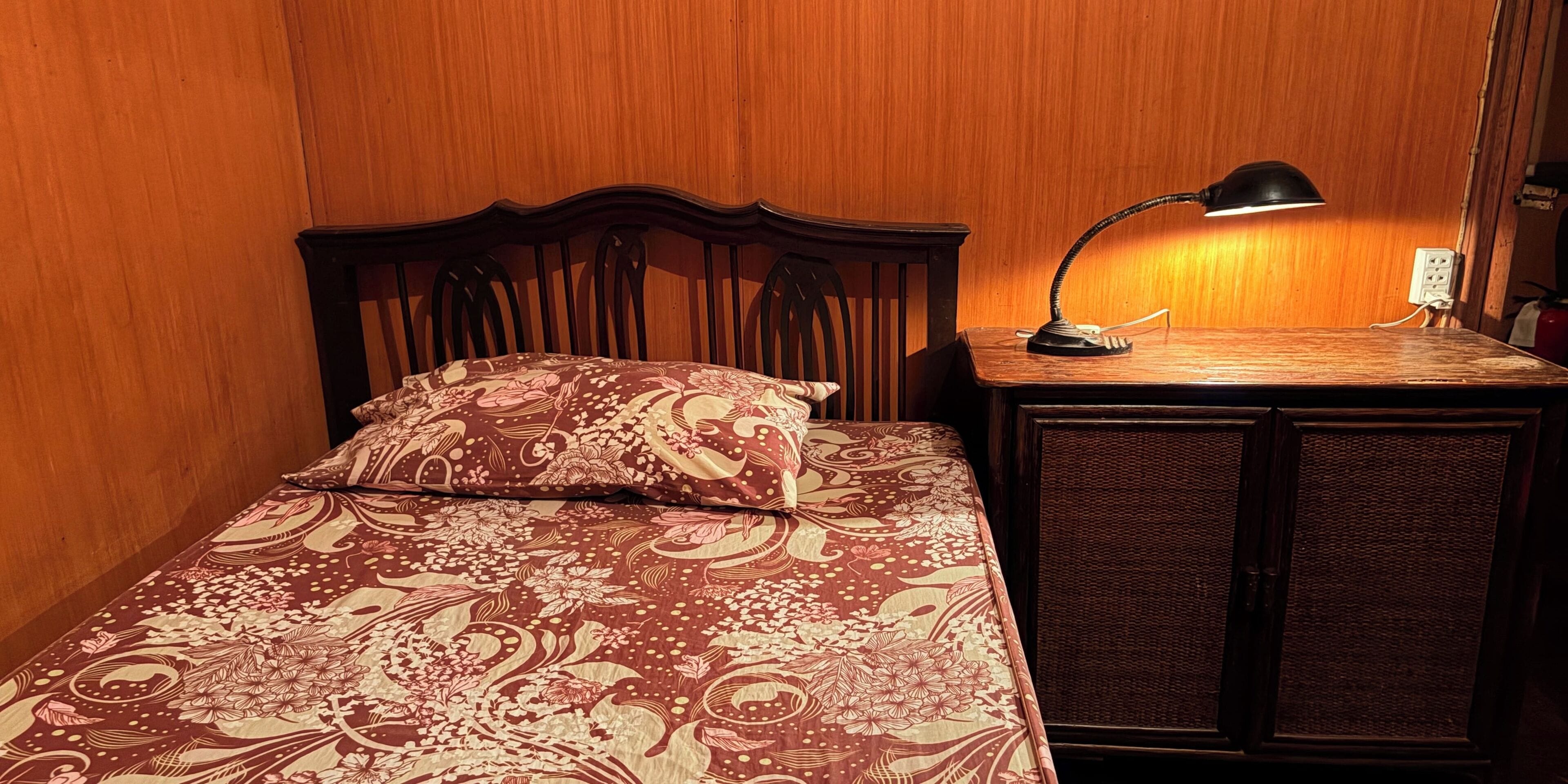 Una cama con cubrecama estampado con motivos florales, cabecera de madera, mesita de noche con lámpara y una mesita auxiliar de mimbre.