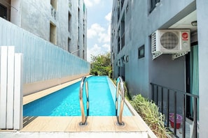 A narrow swimming pool with metal railings, surrounded by buildings with balconies and an air conditioning unit on the right.