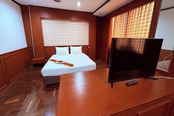 A hotel room with a wooden floor, a bed with white linens, a wooden desk with a remote control, and a window with patterned curtains. - G2 Ayutthaya (Uthai)