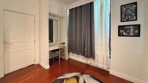 Une pièce avec une porte blanche, une coiffeuse avec un miroir, une chaise, une grande fenêtre avec des rideaux et deux tableaux encadrés accrochés au mur.