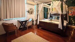 Une chambre à coucher avec un lit à baldaquin, un coin salon avec un fauteuil et une table, et une grande fenêtre avec des rideaux.
