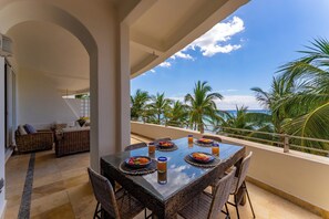 Un comedor con mesa de vidrio y sillas de mimbre, con vistas a un balcón bordeado de palmeras y vista despejada al océano.