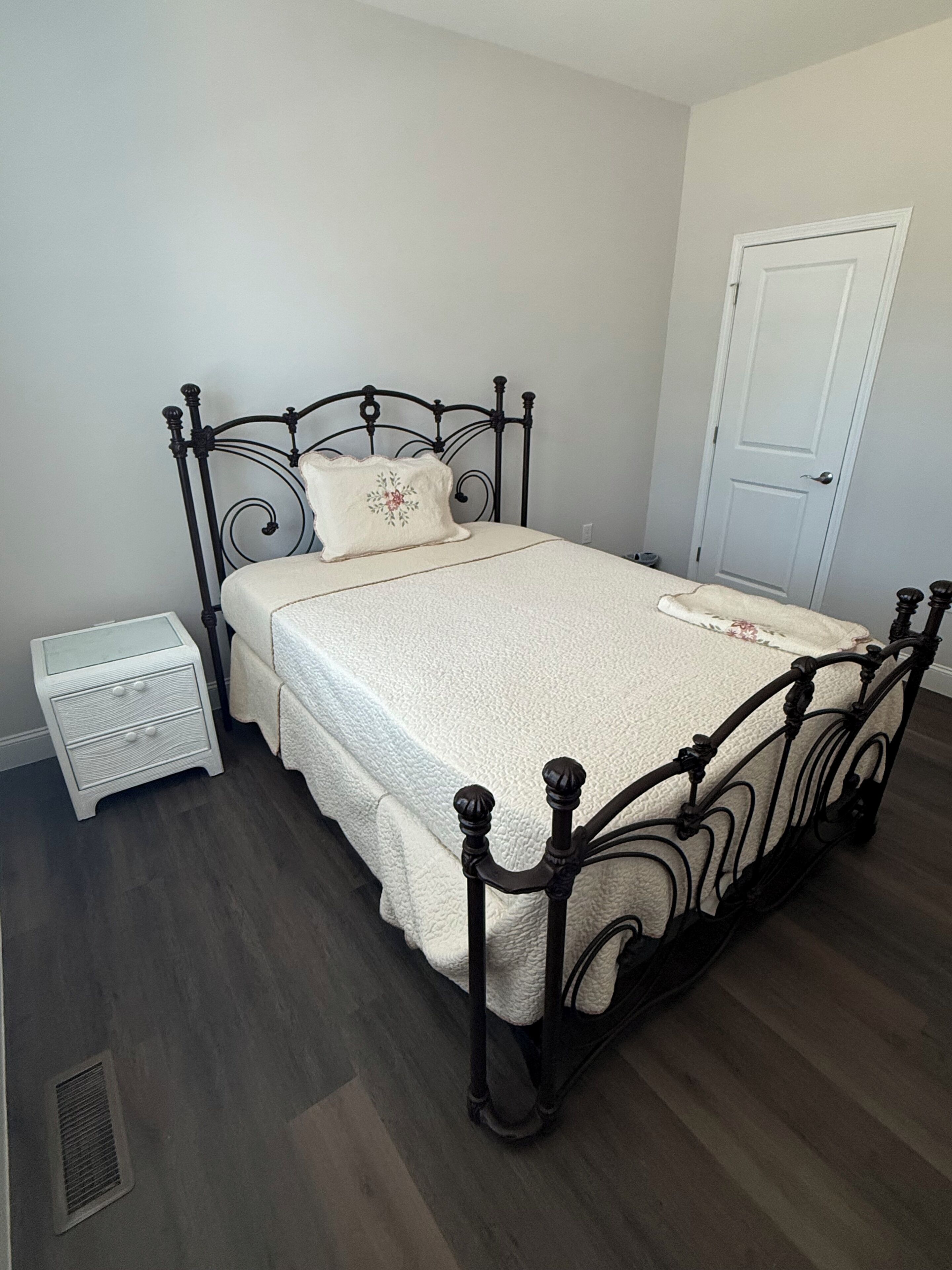 Een net opgemaakt bed met een witte dekbedovertrek en een decoratief kussen, een wit nachtkastje met lades, en een witte deur in een kamer met houten vloeren.