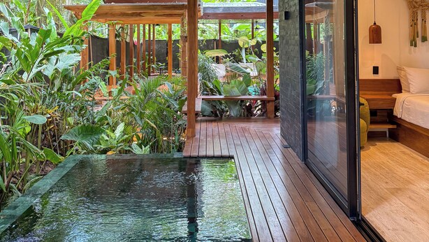 A wooden deck with a small pool, surrounded by lush greenery and a glass door leading to a bedroom. - Kaia Bungalow – Adults Only Private Pool (Manzanillo)