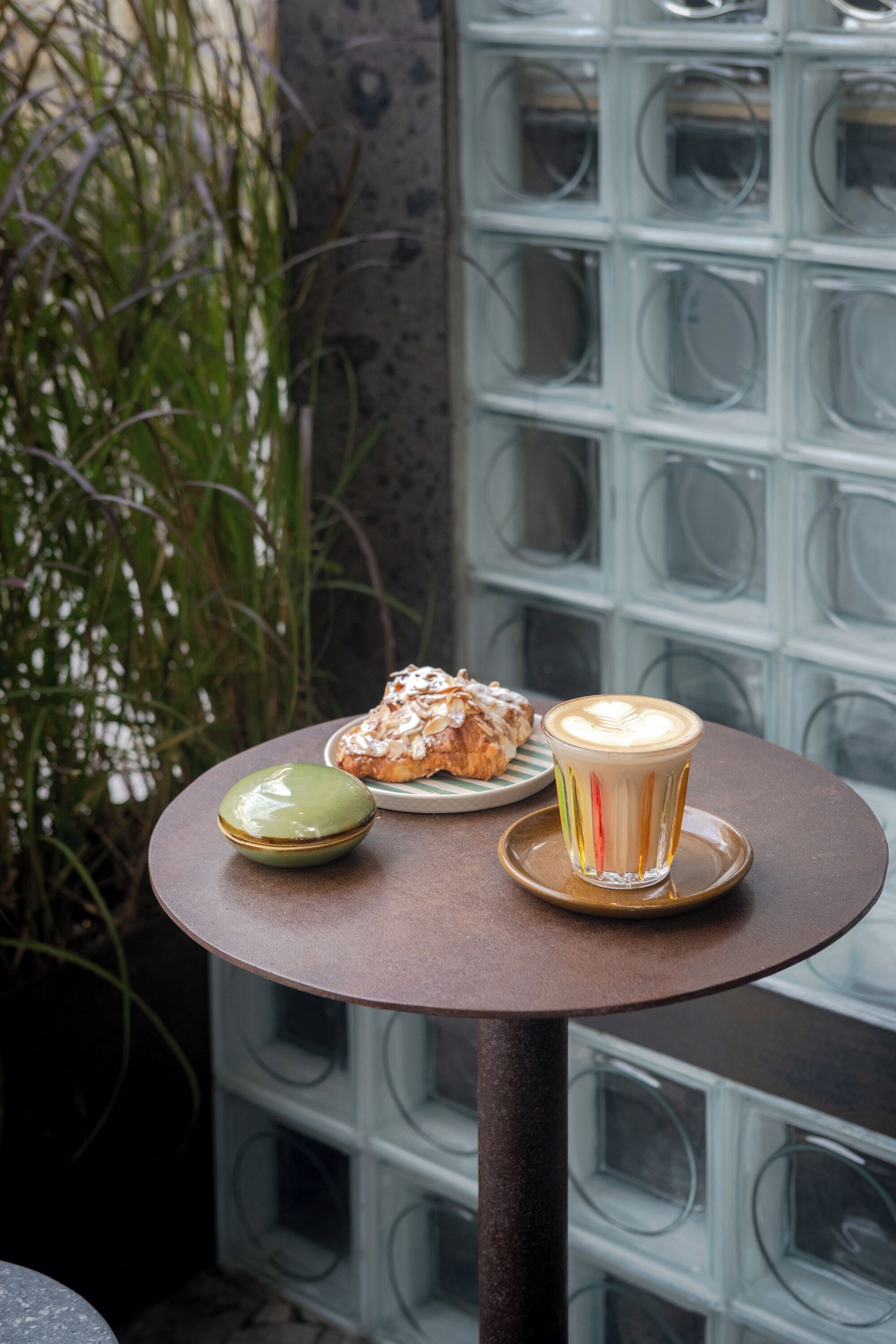 A table with a pastry and a cup of coffee, set against a backdrop of a patterned wall and greenery.