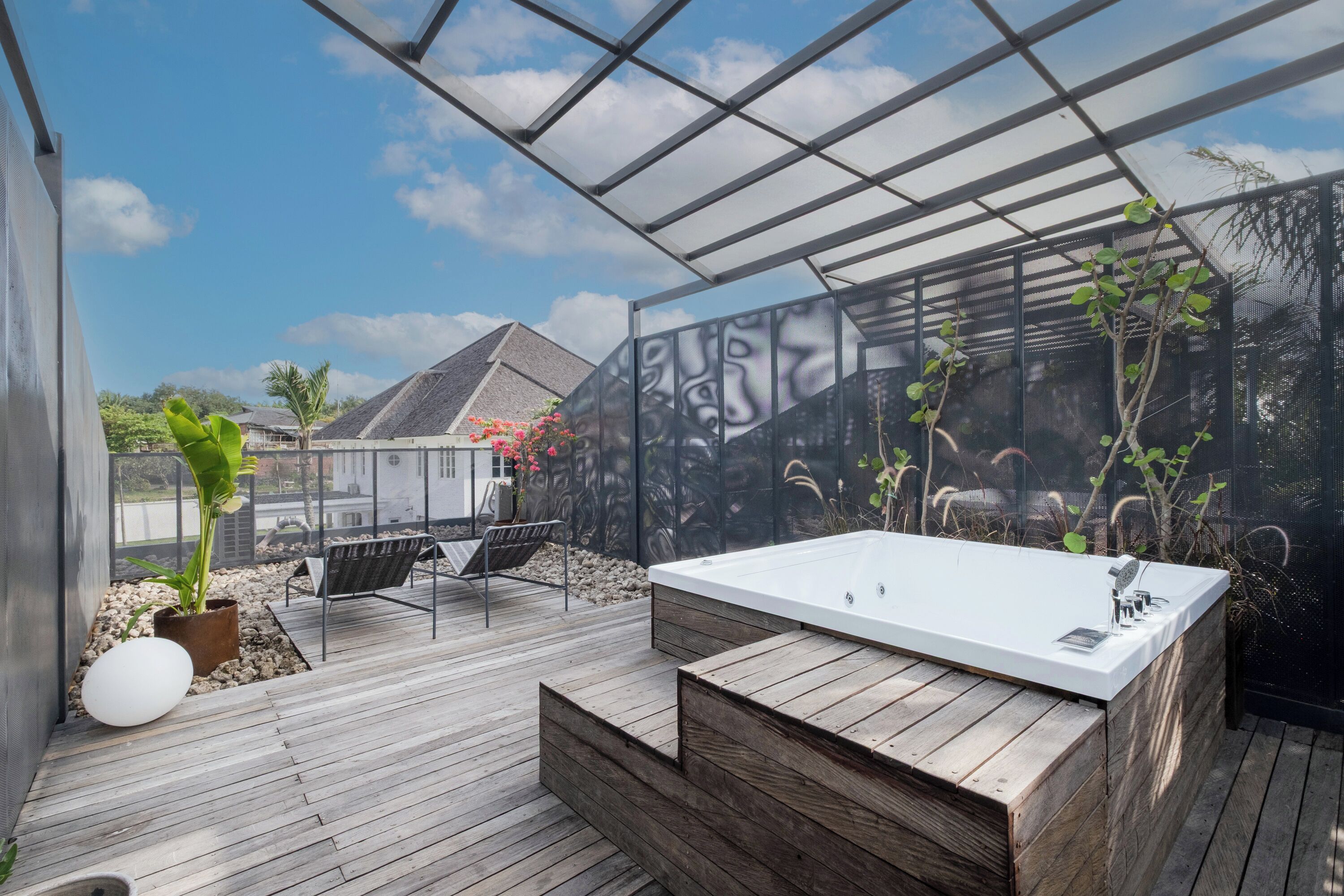 A modern outdoor bathroom with a wooden deck, a large bathtub, and a view of a house and palm trees.